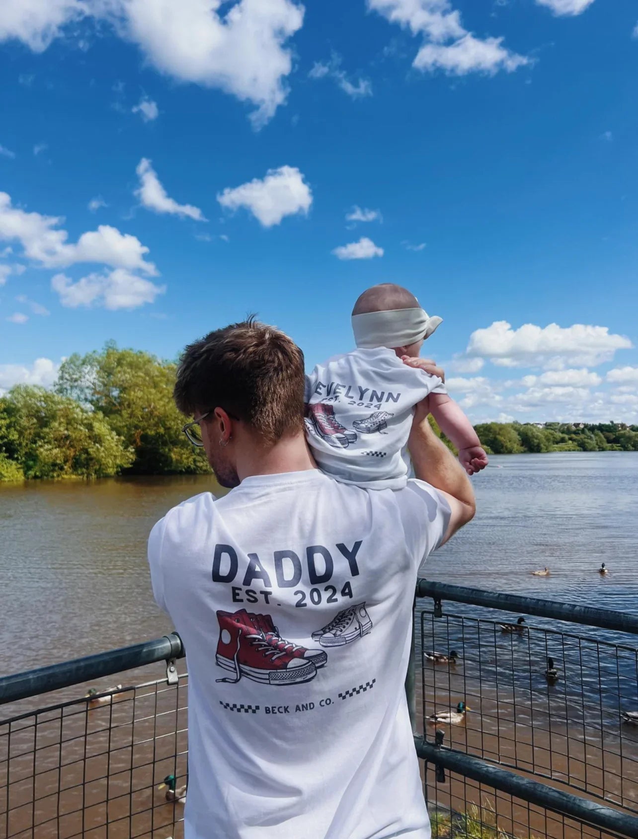 Father and baby in Beck and Co. matching streetwear tees by a lake, blue sky background