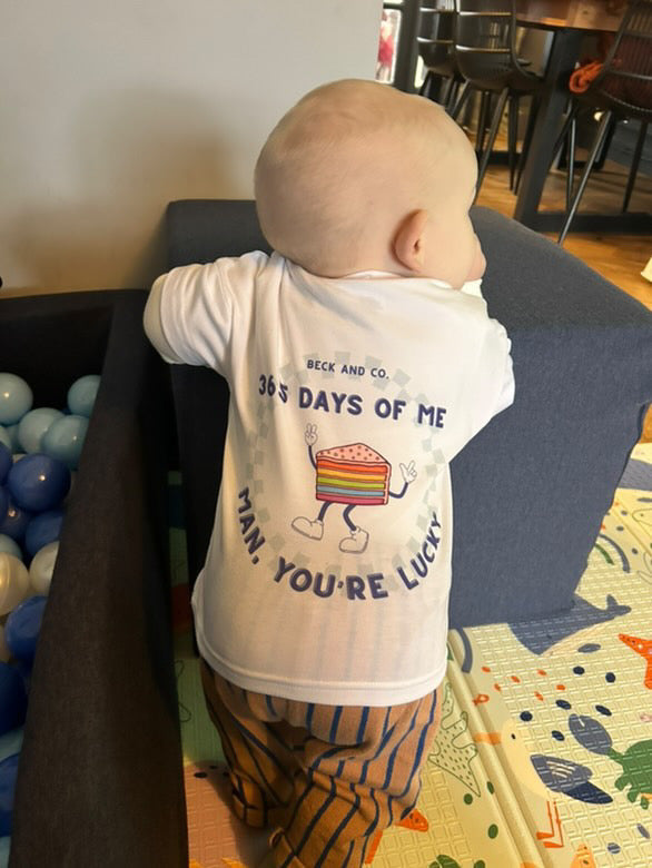 Baby wearing a shirt with text and graphics, standing in a room with furniture and toys.