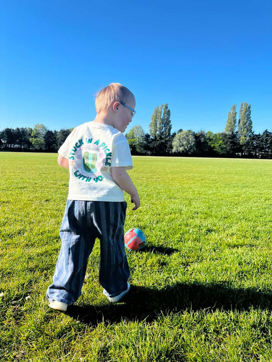 Child in graphic oversized t-shirt playing with a soccer ball on a grassy field with a clear blue sky.