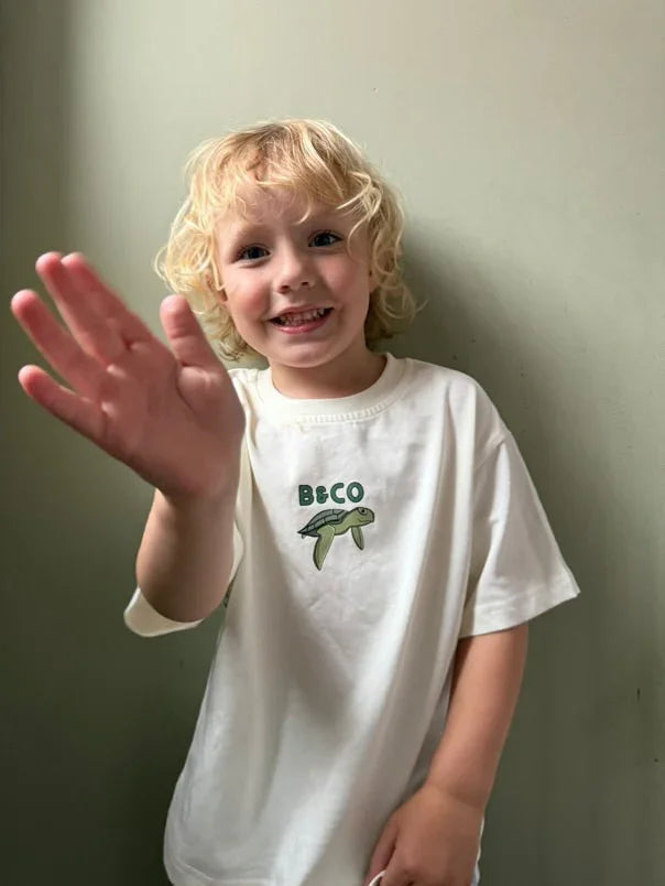 Child wearing a white t-shirt with a logo, waving and smiling against a plain background