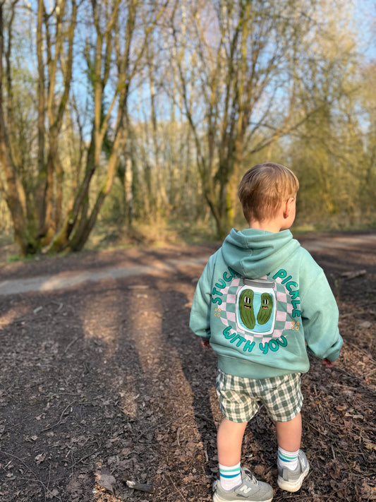Child wearing a hoodie and lightweight gingham shorts with a design and text, standing on a path in a forest.