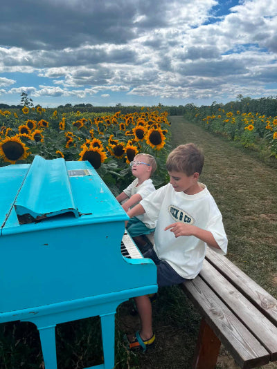 Two boys sitting on a wooden bench next to a bright turquoise piano in a sunflower field under a cloudy sky.