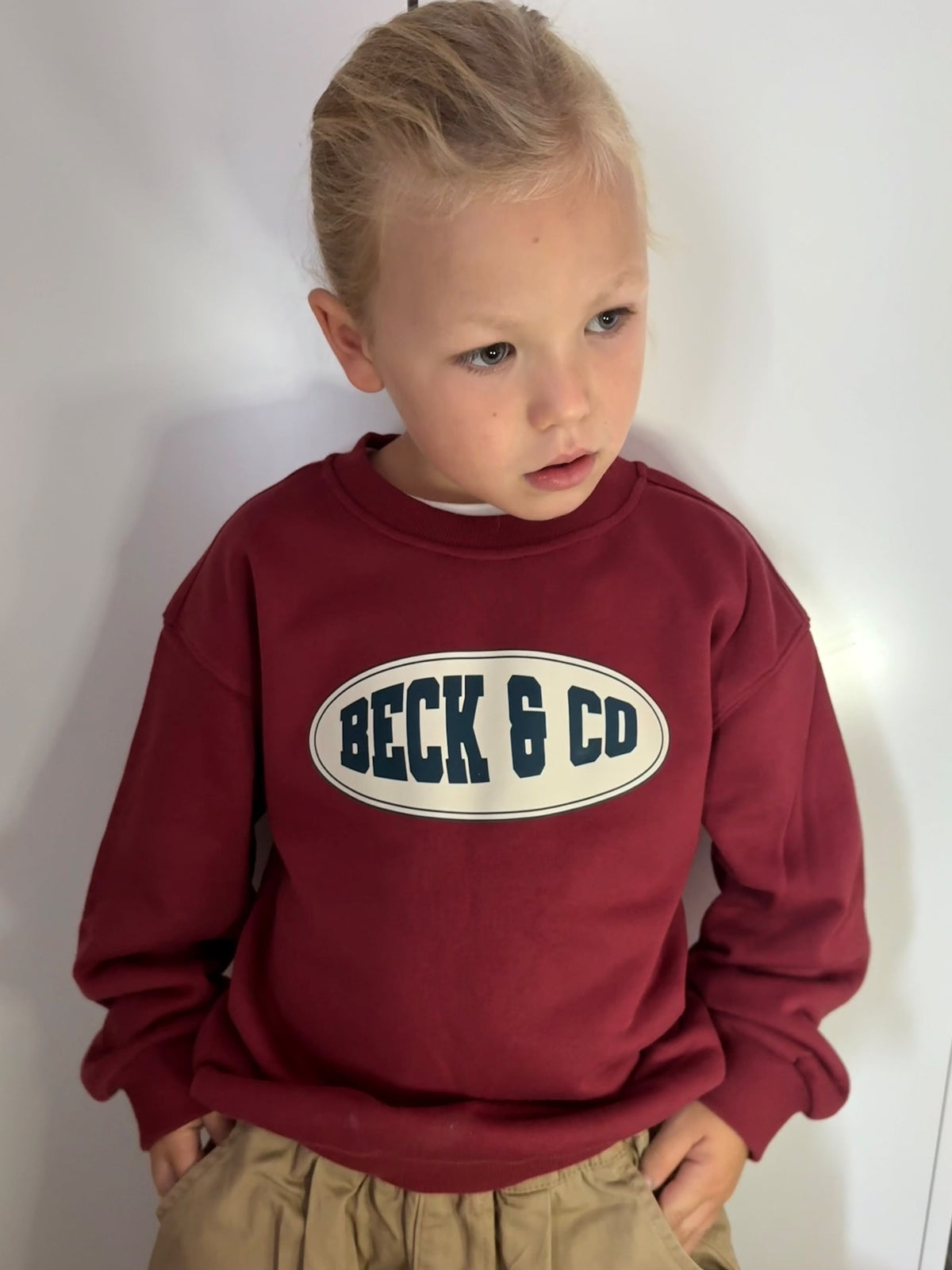 A young child wearing a burgundy crewneck sweatshirt with an oval 'BECK & CO' logo on the chest, paired with khaki pants, standing against a plain white wall.