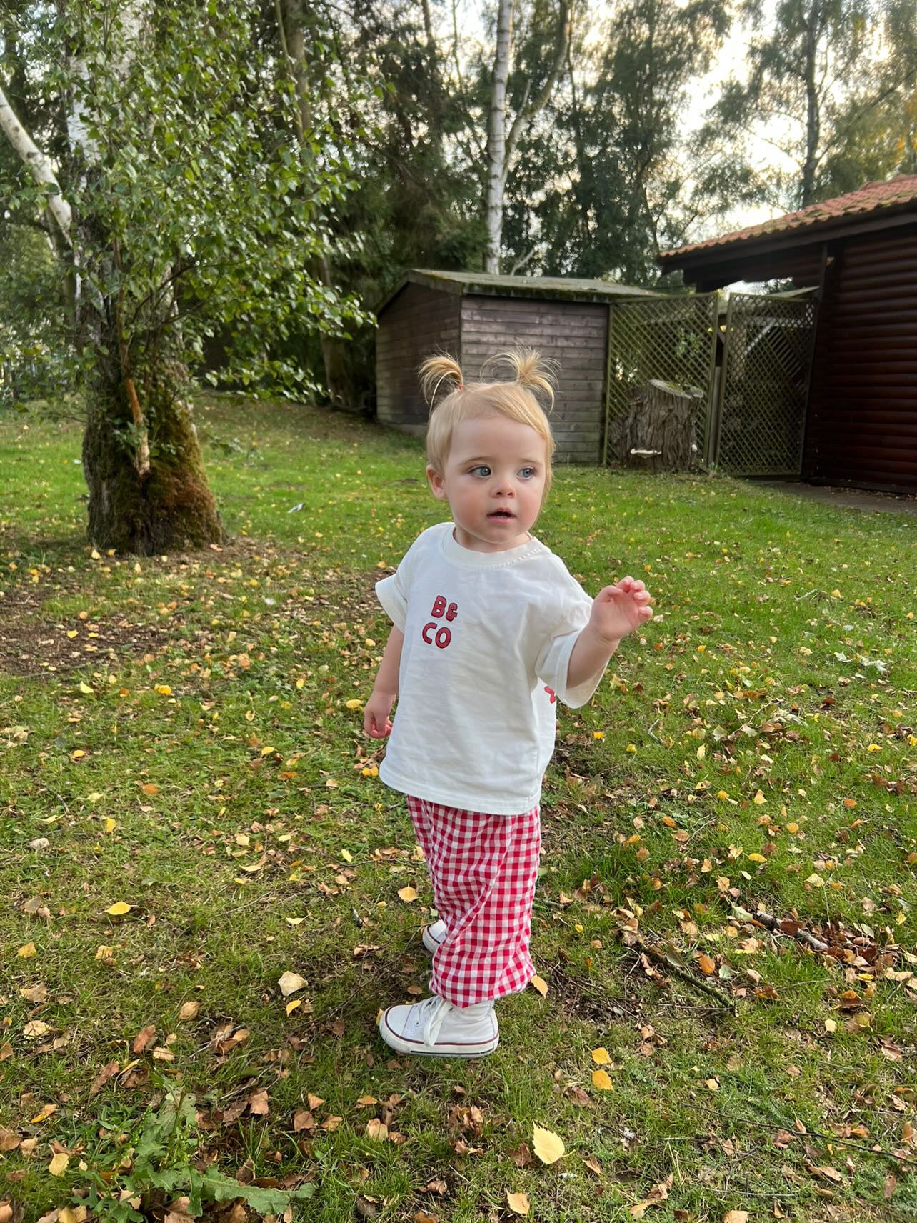 Child in a white shirt and red checkered pants standing in a grassy area with trees and wooden structures in the background.