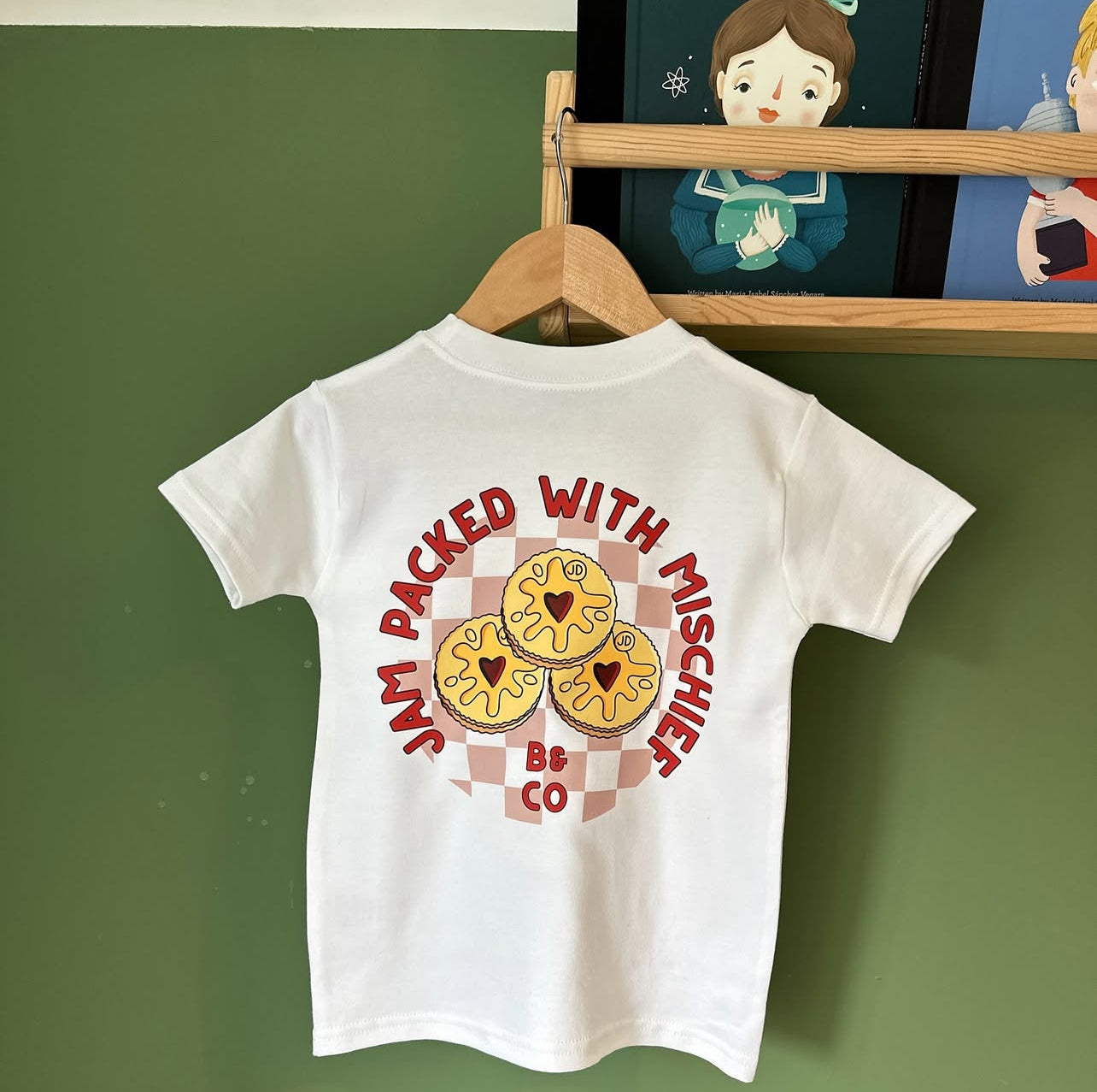 White t-shirt with cookie design and text on a hanger against a green wall.