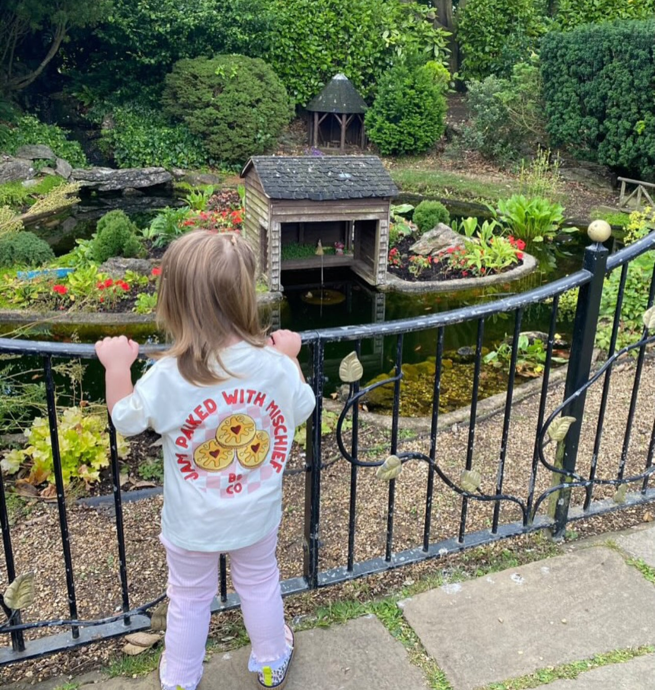 Child wearing a shirt with a design, standing by a garden fence overlooking a pond and garden.