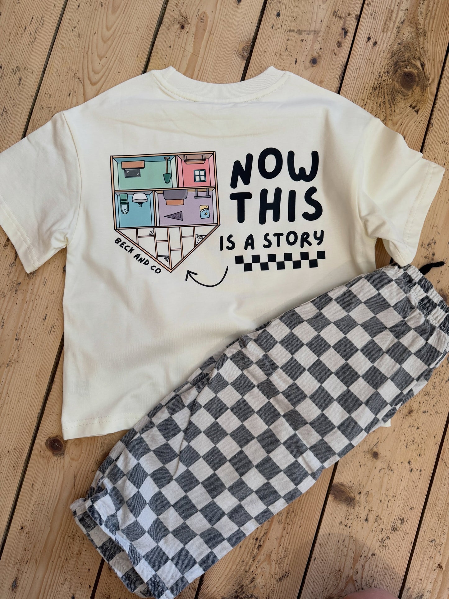 A children's outfit laid flat on a wooden floor, featuring a cream short-sleeve T-shirt with a graphic that reads 'NOW THIS IS A STORY' and a small house illustration, paired with black-and-white checkered pants.