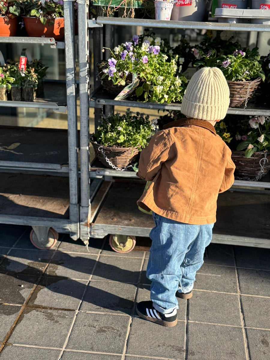 Child in a brown jacket and white beanie standing next to a cart filled with plants at a nursery.