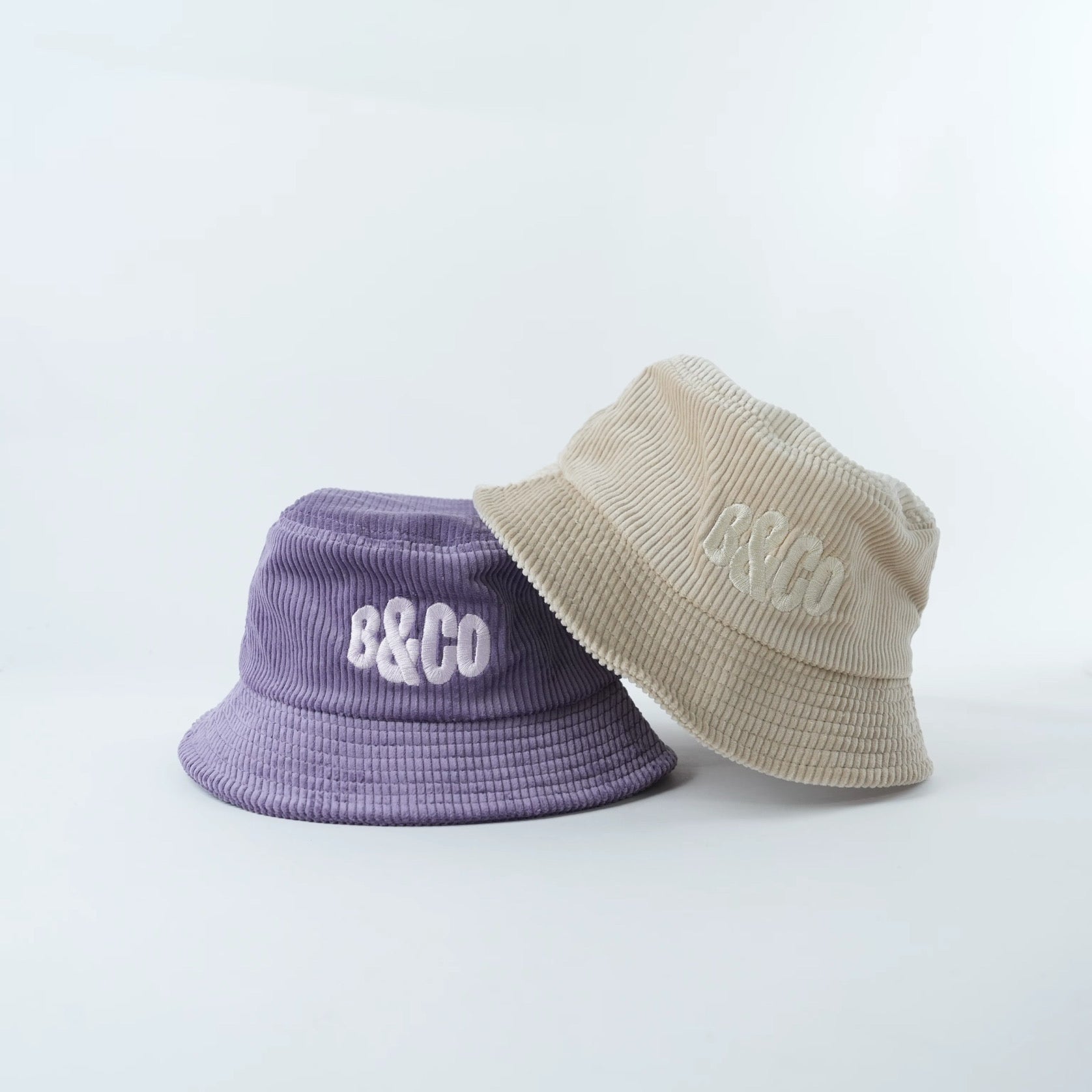 Two bucket hats, one purple and one beige, with 'B&Co' branding on a light gray background.