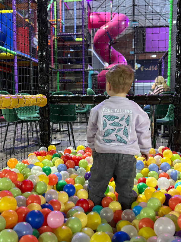 Child wearing oversized butterfly illustrated sweatshirt playing in a ball pit at an indoor playground with colorful equipment in the background.