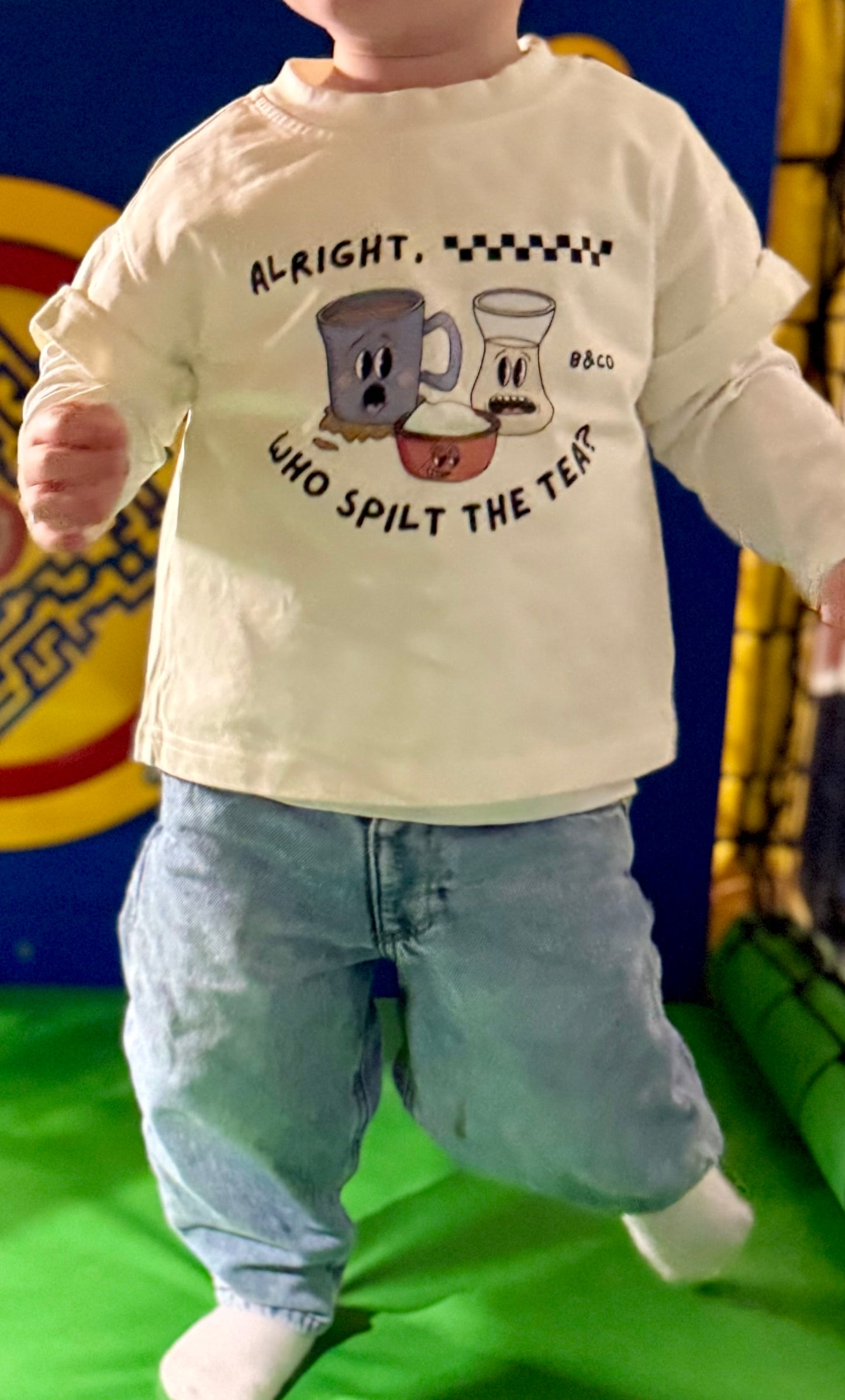Child wearing a white t-shirt with a graphic design and text, standing on a green surface with a colorful background.