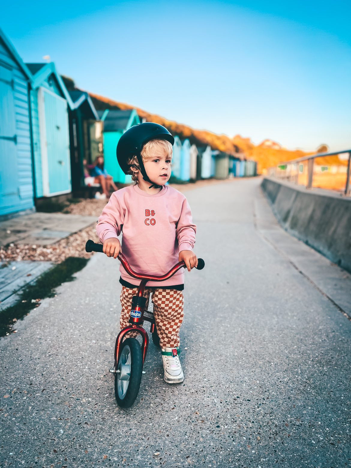 Child riding a balance bike on a path with beach huts in the background wearing a pink skater style sweatshirt