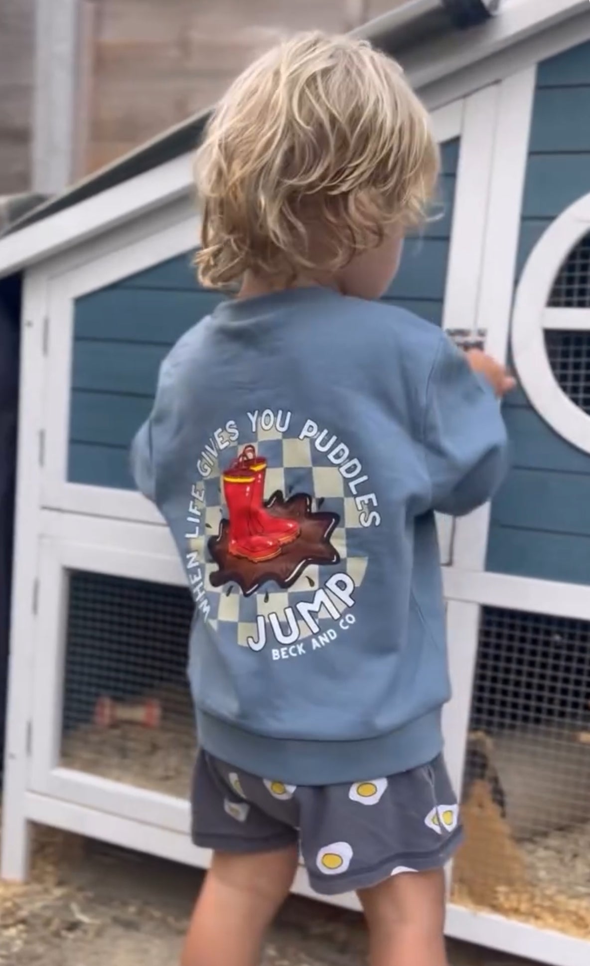 Child wearing a blue sweatshirt with a graphic design in front of a chicken coop.