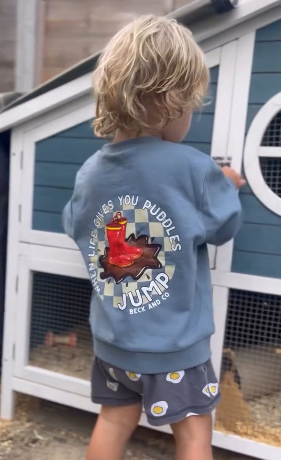 Child wearing a blue sweatshirt with a graphic design in front of a chicken coop.