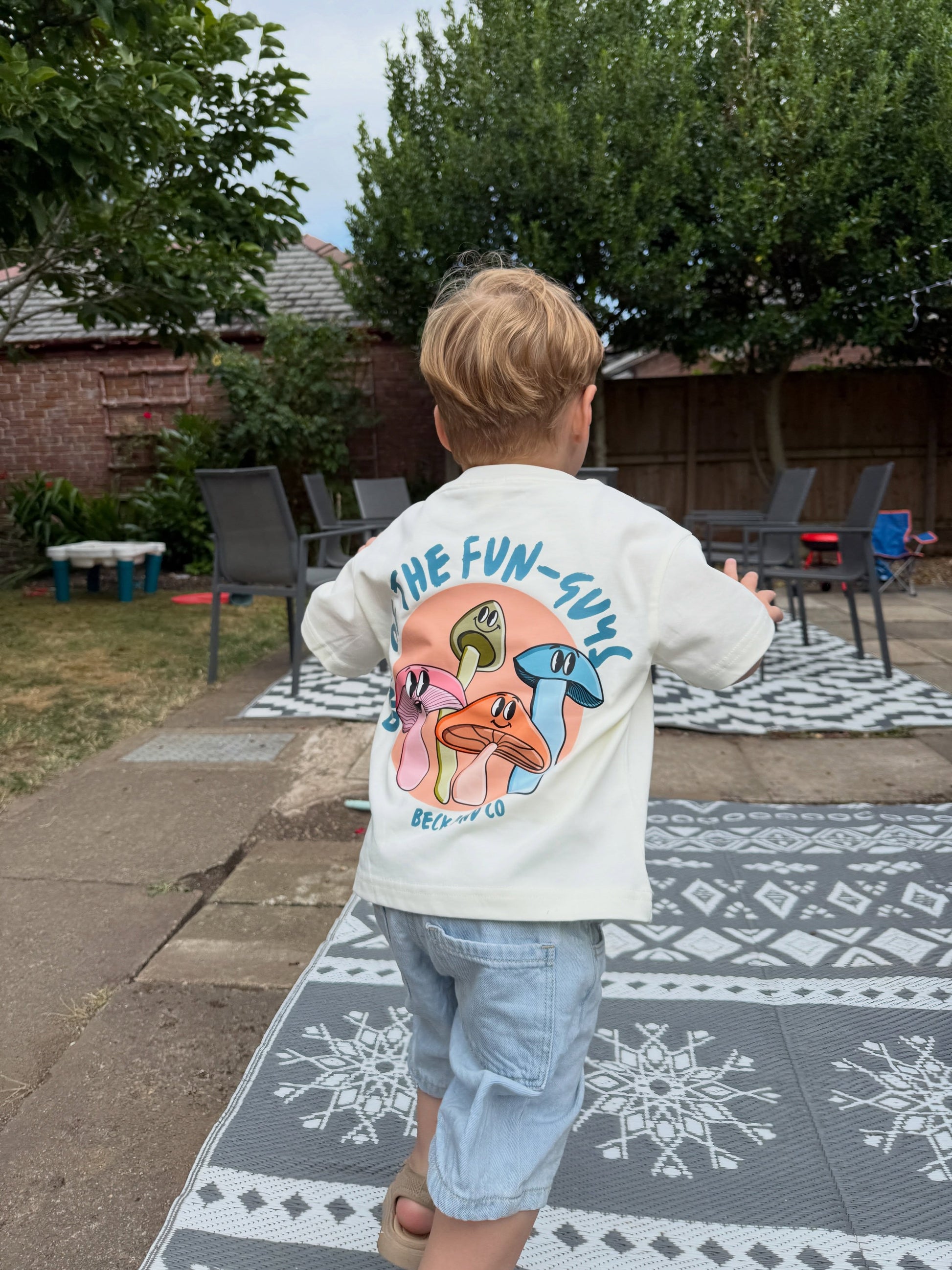 A child viewed from behind wearing a cream-colored t-shirt with a colorful mushroom graphic and the text 'ONE OF THE FUN-GUYS' on the back, paired with light blue denim shorts in an outdoor backyard setting.