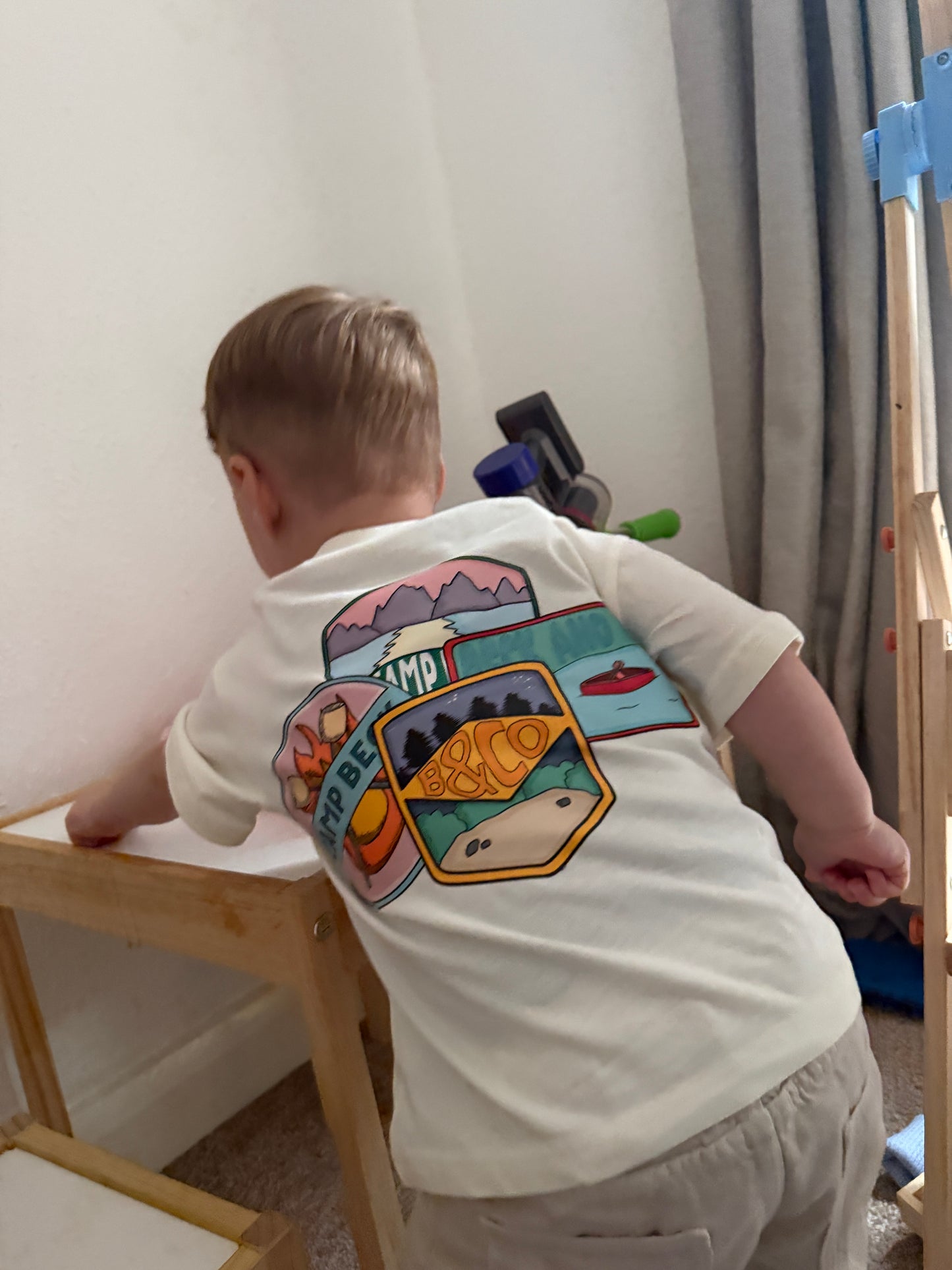 Person wearing a white t-shirt with colorful camping graphic design, standing on a wooden stool.