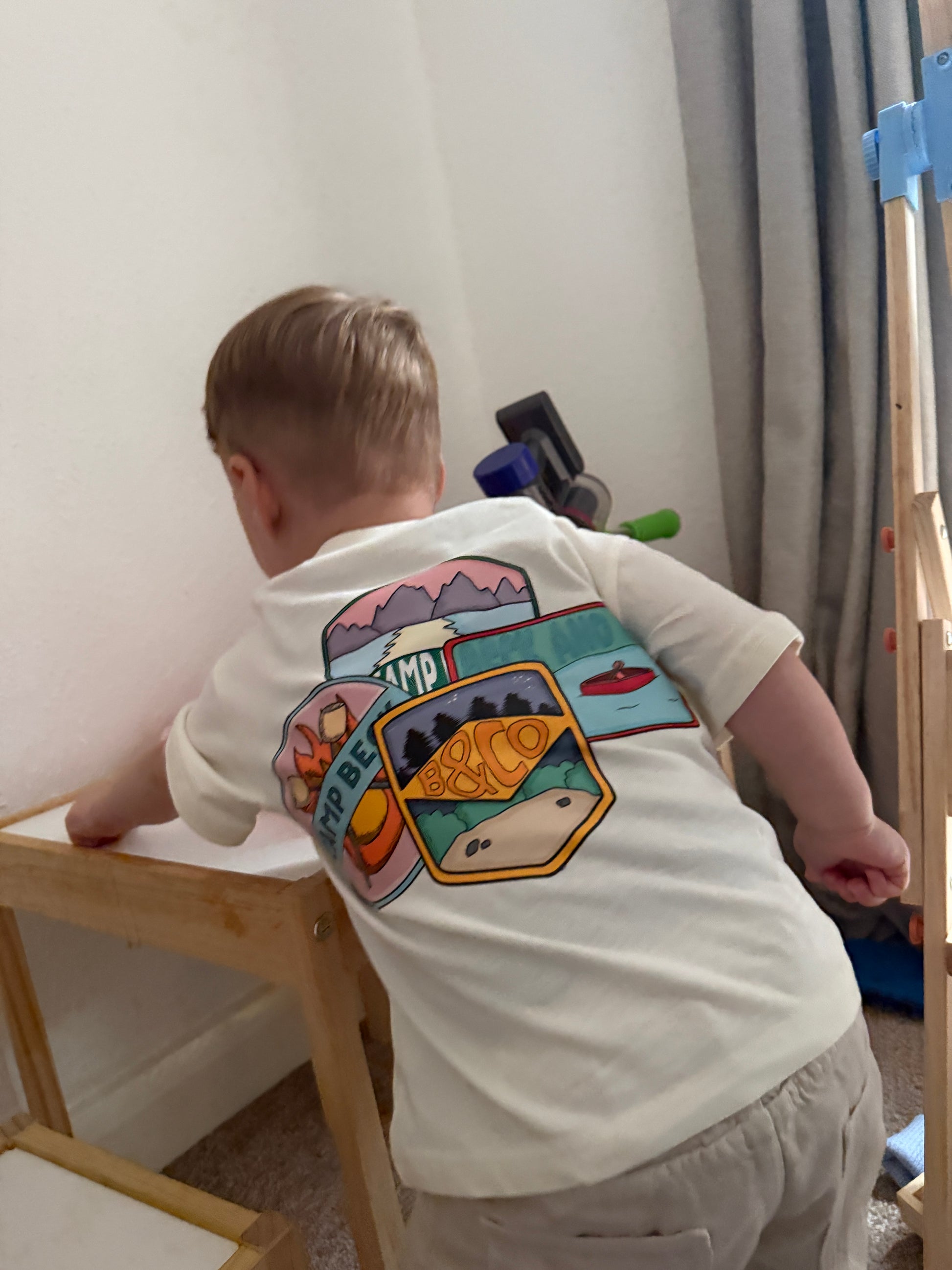 Person wearing a white t-shirt with colorful camping graphic design, standing on a wooden stool.