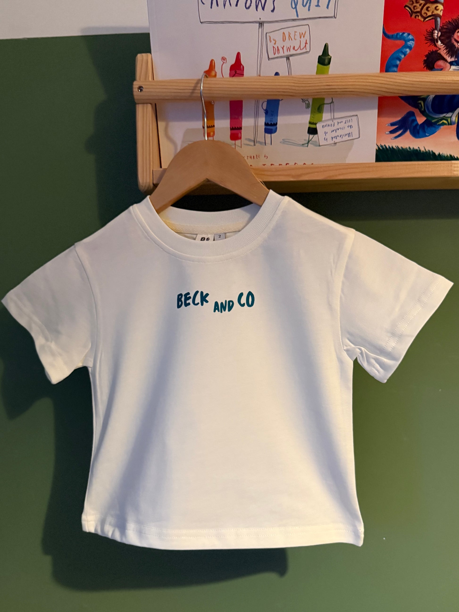 White t-shirt with 'BECK AND CO' text on a hanger against a green wall.
