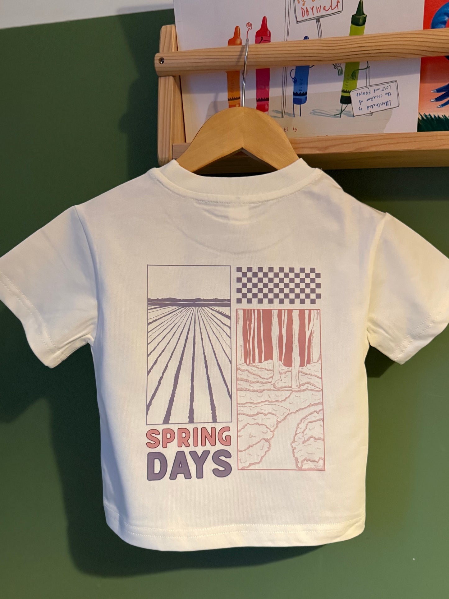 White t-shirt with 'Spring Days' design on a hanger against a green wall.