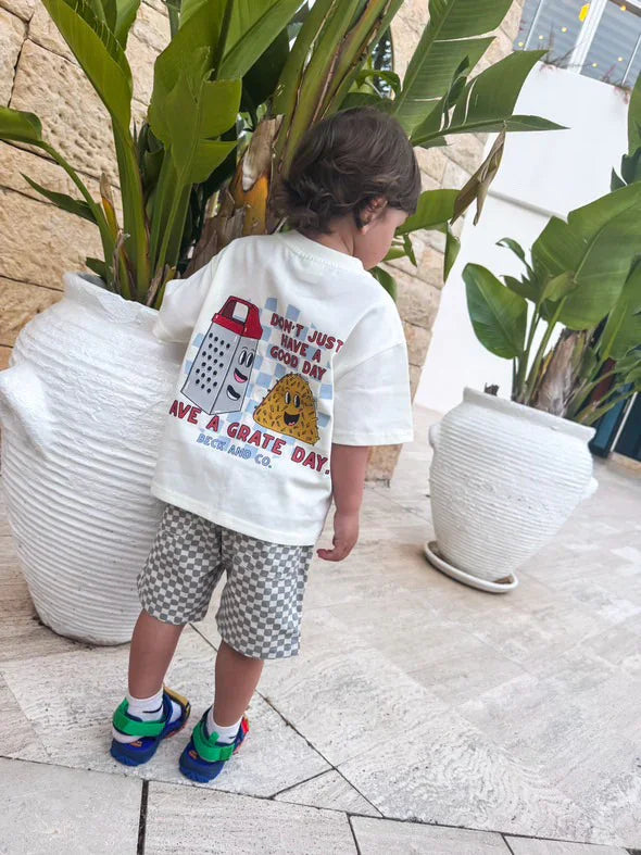 Child wearing Beck and Co. graphic tee and checkered shorts outdoors near large potted plants