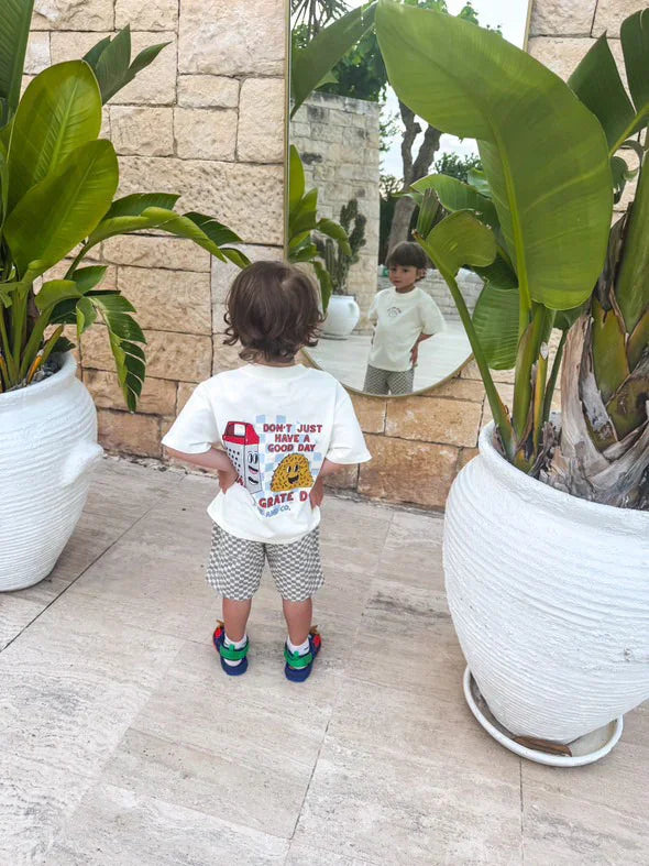 Young child in Beck and Co. streetwear graphic tee stands between potted plants, looking into a mirror outdoors.