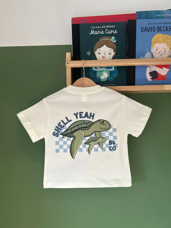 Children's white t-shirt with turtle graphic and text on a green surface with books in the background