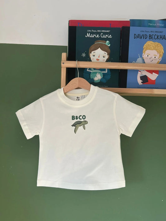 White t-shirt with a turtle design on a hanger against a green wall with books in the background