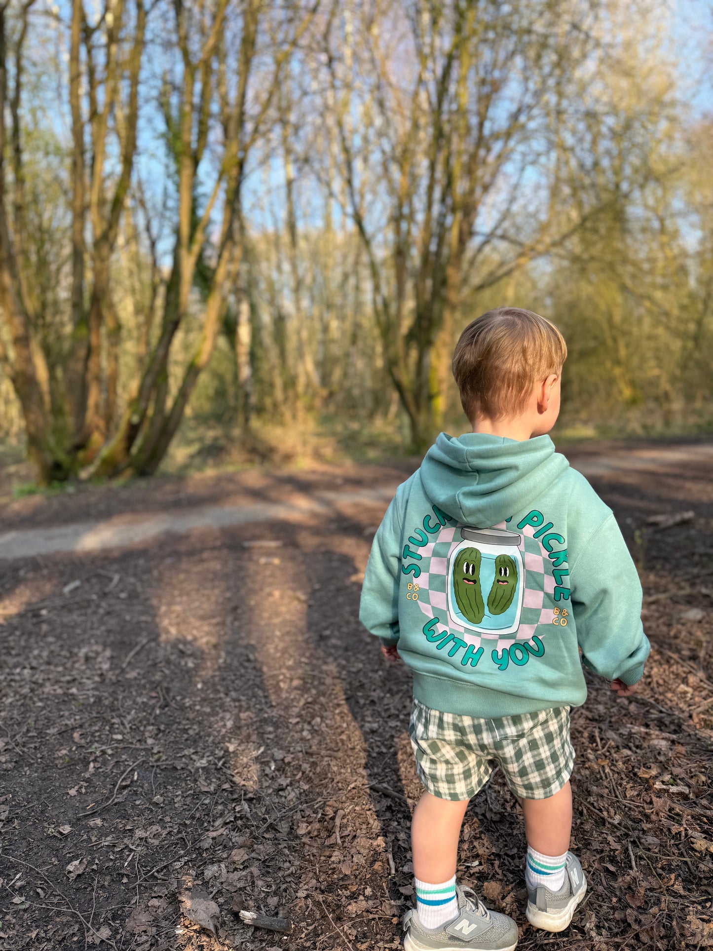 Child wearing a hoodie and lightweight gingham shorts with a design and text, standing on a path in a forest.
