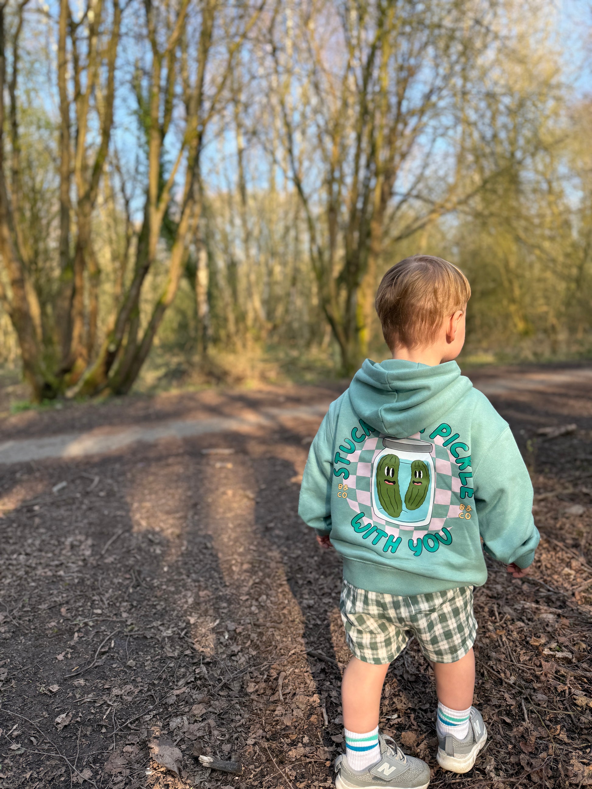 Child wearing a hoodie and lightweight gingham shorts with a design and text, standing on a path in a forest.