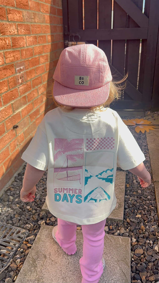 Child wearing a white t-shirt with 'Summer Days' text and a pink cap, standing on a stone path.
