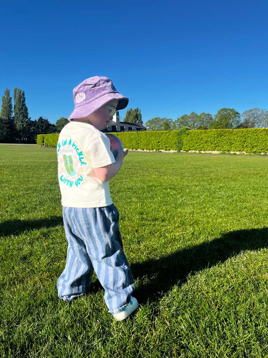 Child wearing a purple hat and white graphic t-shirt with blue stripes standing on grass