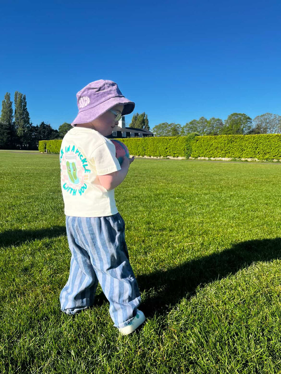 Child wearing a purple hat and white graphic t-shirt with blue stripes standing on grass