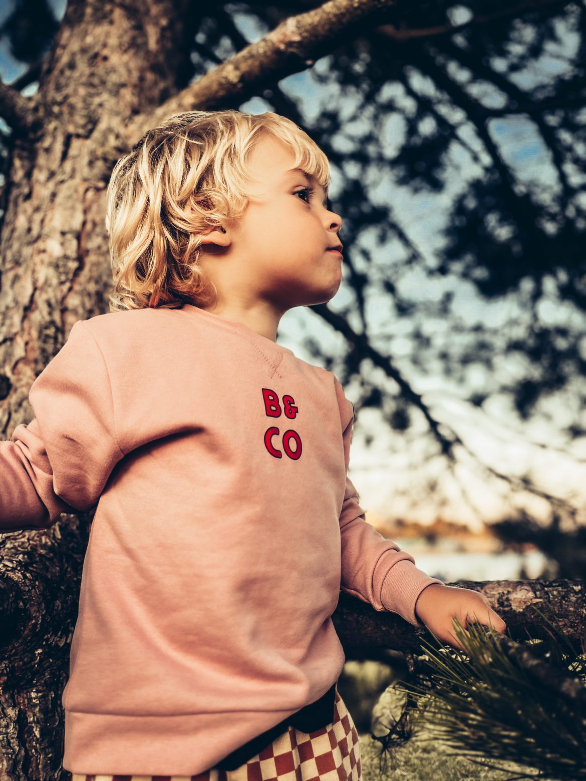Child wearing a pink sweatshirt with Beck and Co logo, leaning against a tree in a natural setting.