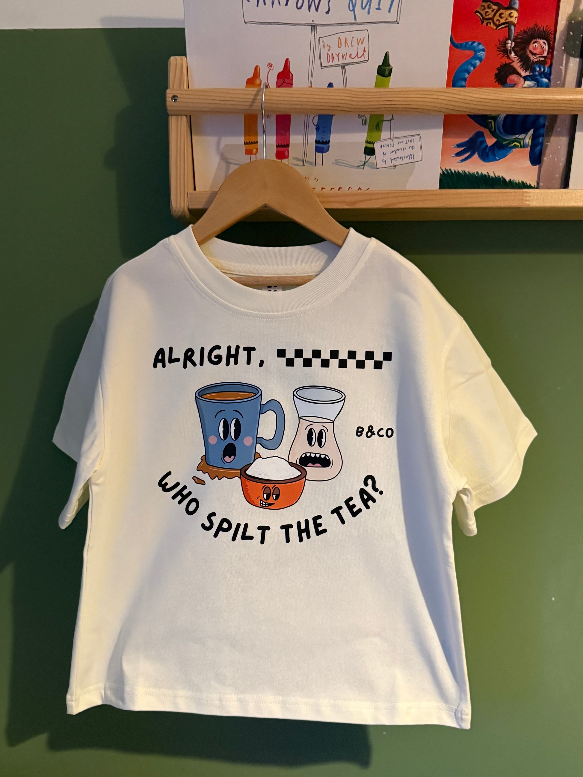 Children's white t-shirt with graphic design and text on a hanger against a green wall.