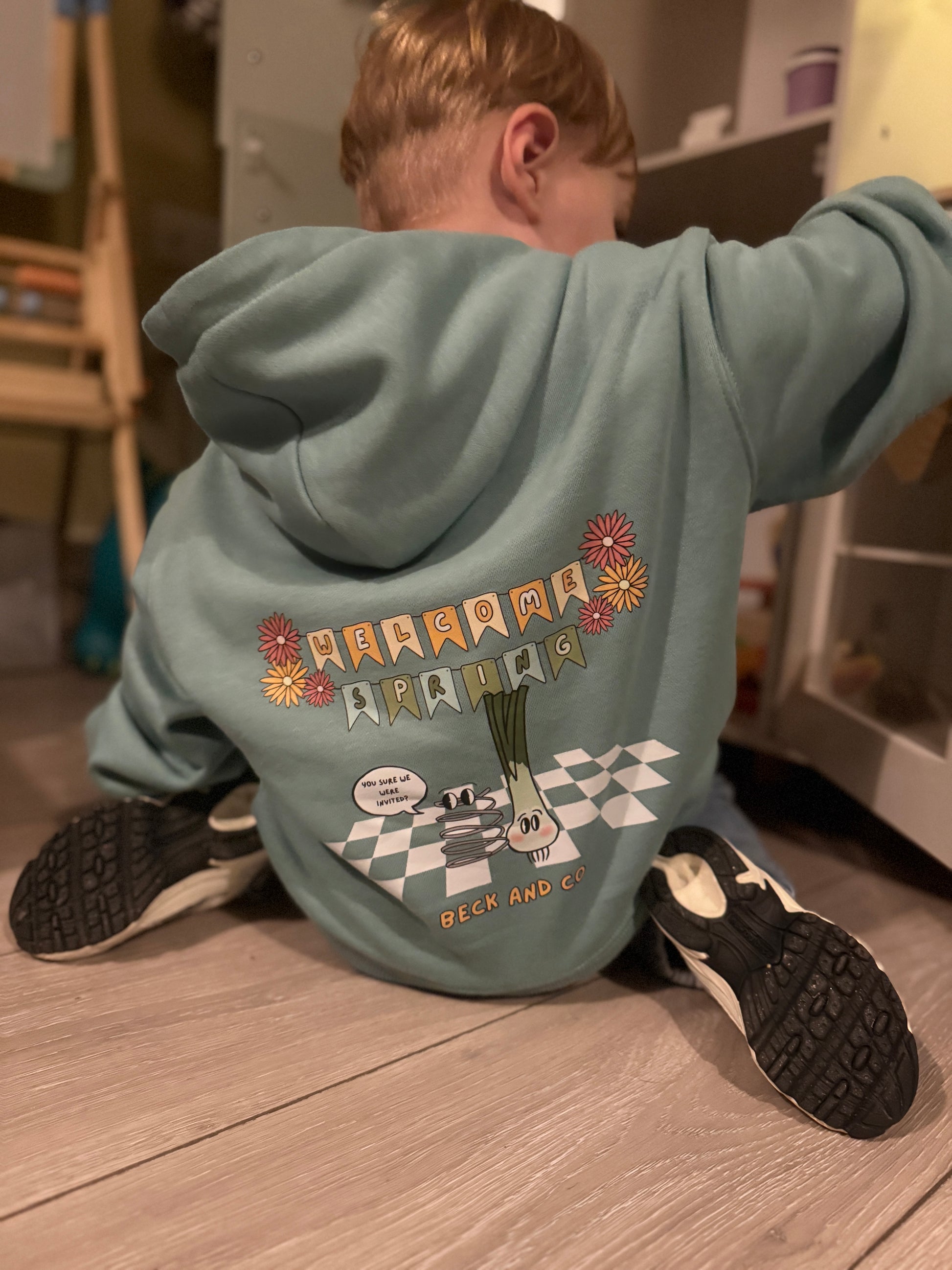 Toddler wearing a graphic hoodie with text and graphics on the back, sitting on the floor.